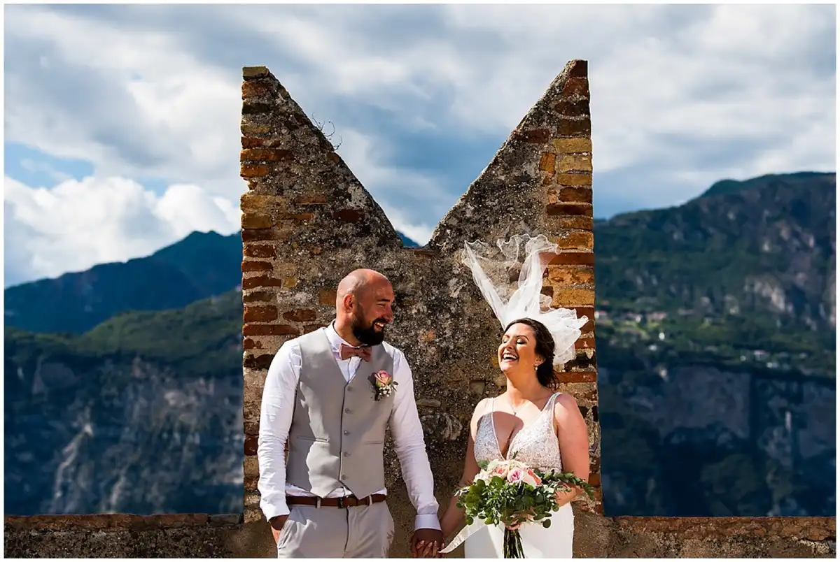 Bride and groom laughing hand in hand in front of an old brick castle wall, her veil blowing up, mountains and clouds behind.