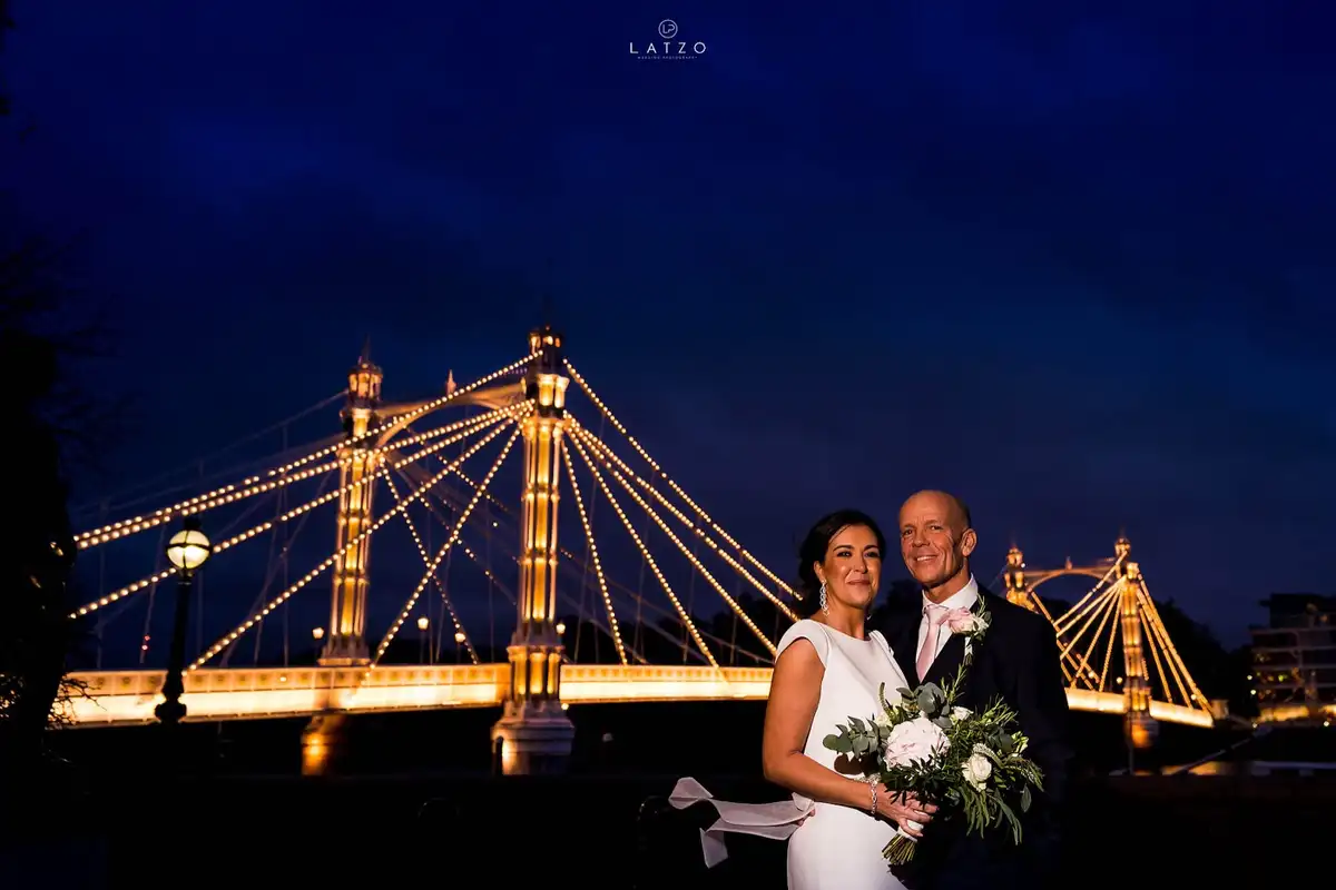 Bride and groom smile at the camera at dusk in front of Albert Bridge, its towers and cables lit up in gold against a dark blue sky.