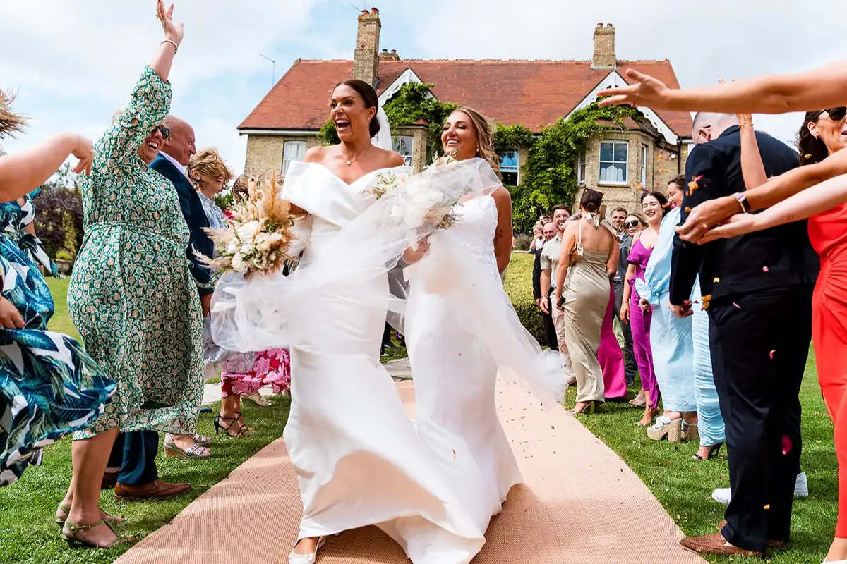 Two brides laugh as they walk up a matting aisle through cheering guests, a veil blowing across them and a brick farmhouse behind.