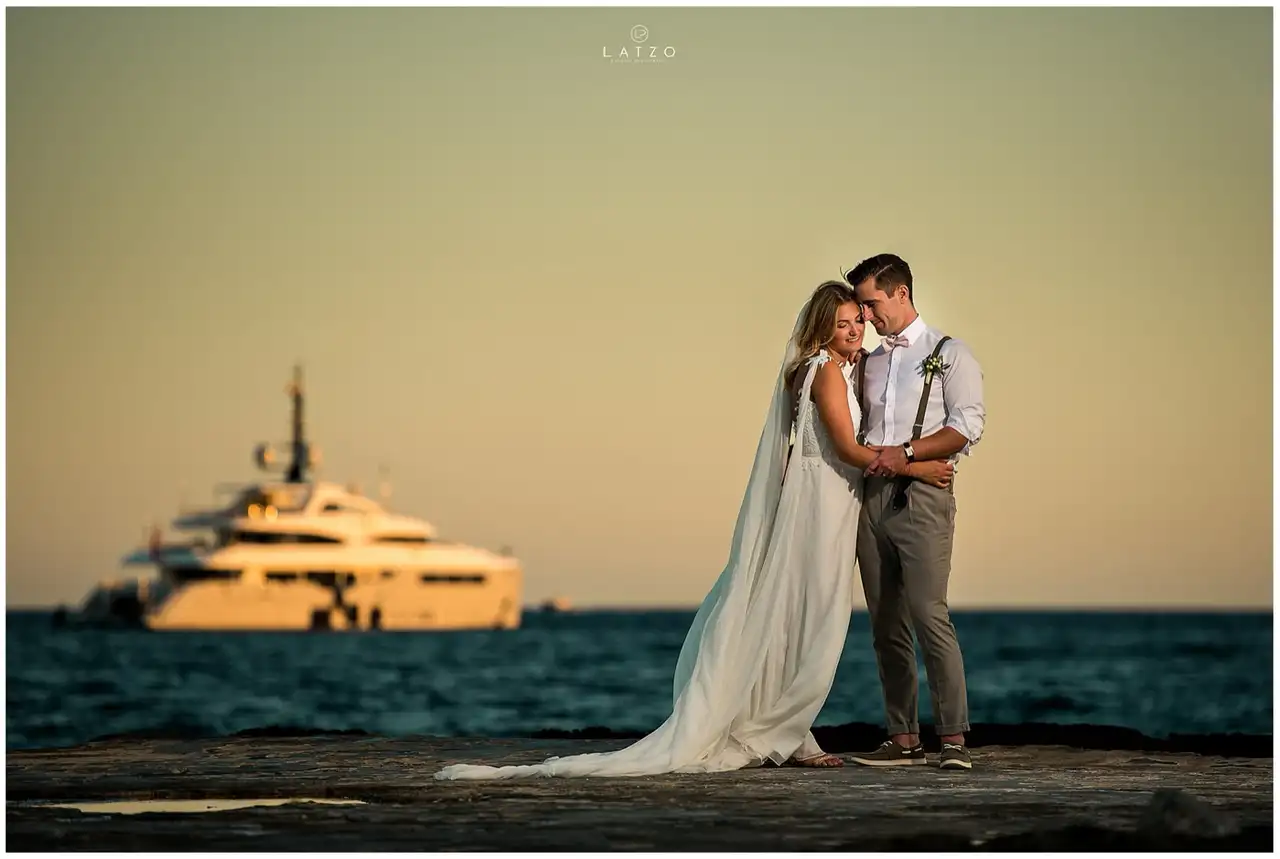 Bride and groom in braces hold each other on a rocky shore at sunset, a large yacht glowing on the sea behind them.