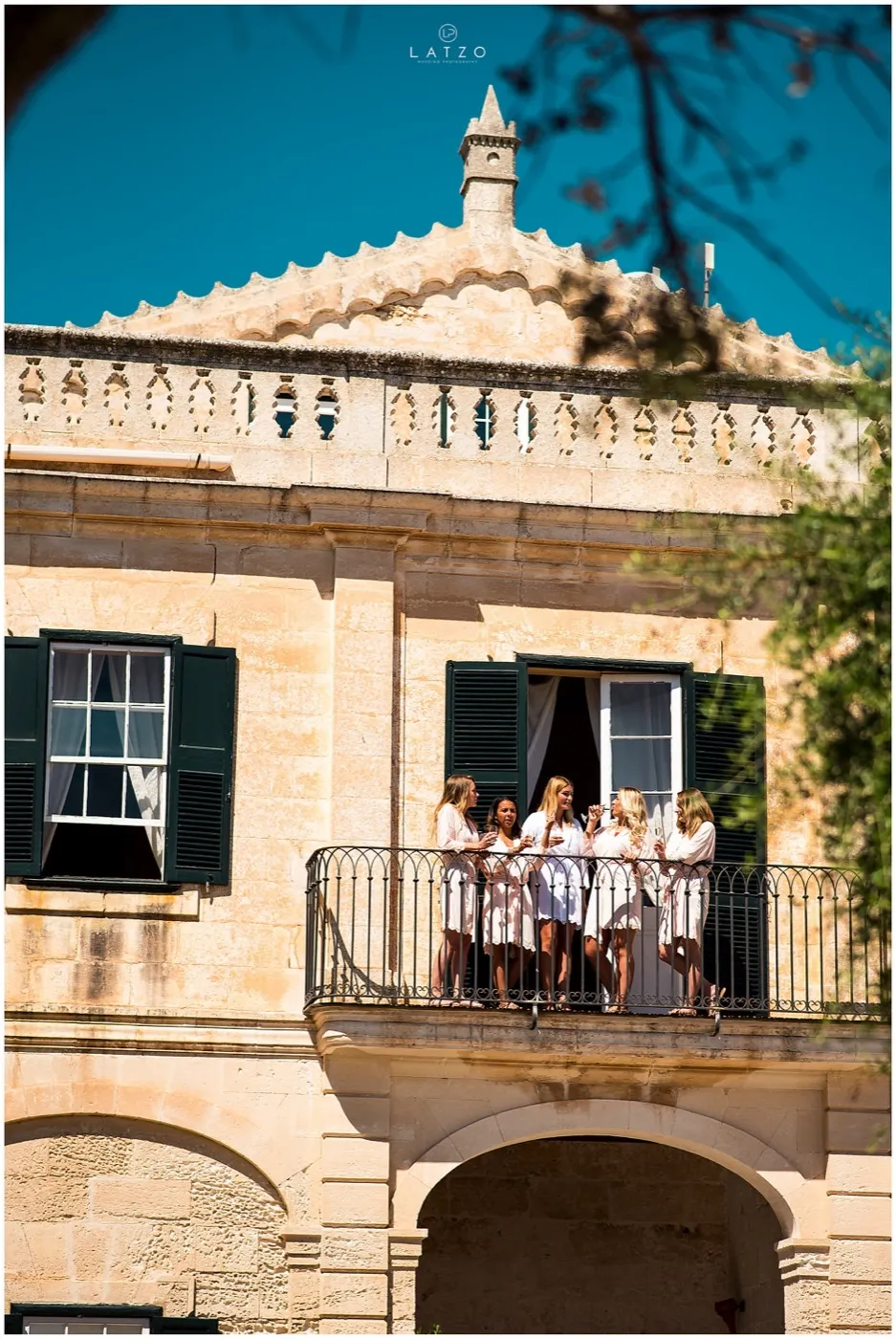 Five women in white robes chat with drinks on the balcony of a sandstone farmhouse with green shutters, under a deep blue sky.