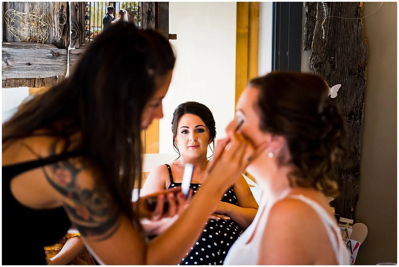 A make-up artist with a tattooed shoulder brushes blusher on to a bride’s cheek while a bridesmaid in a polka dot dress watches in the mirror.