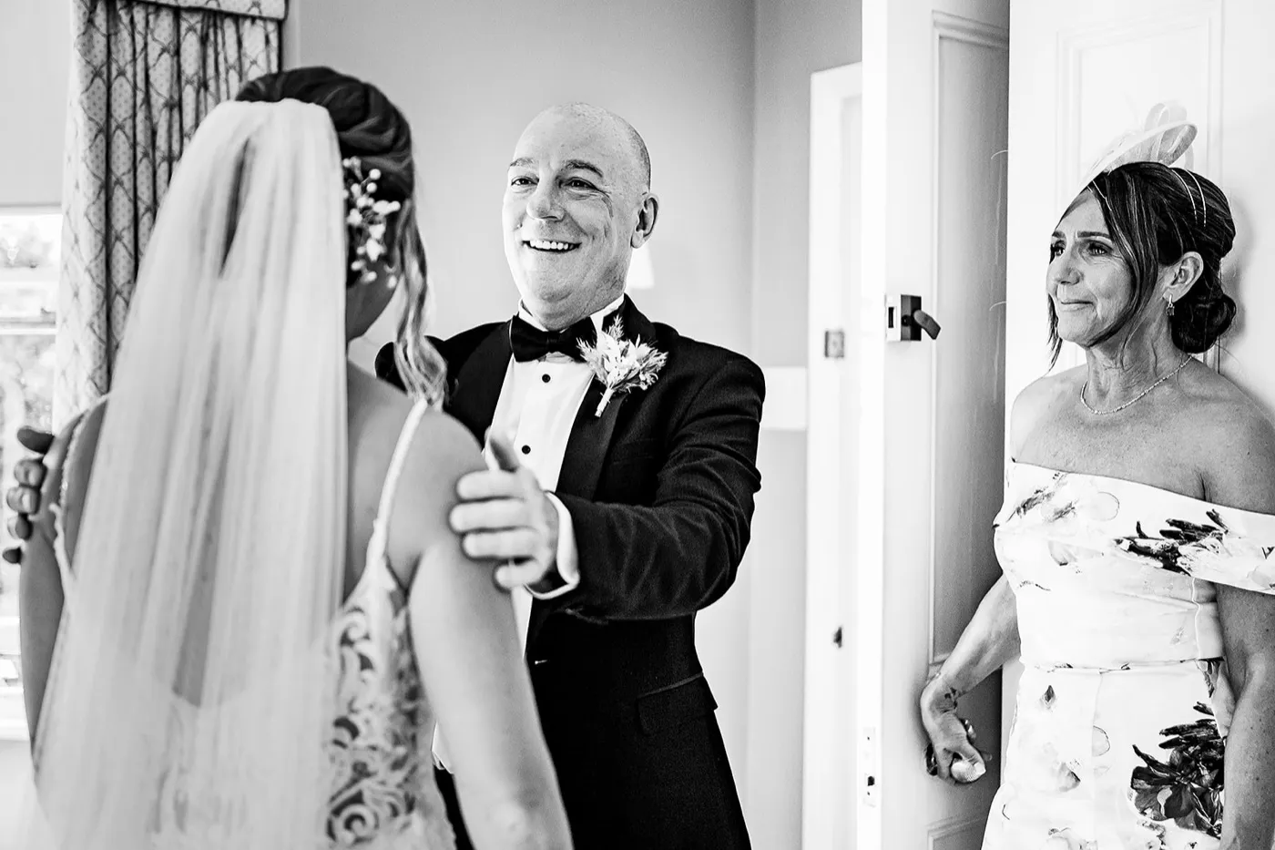 Black and white of a smiling man in a tuxedo with his hands on a bride’s shoulders as a woman in a fascinator watches from the doorway.
