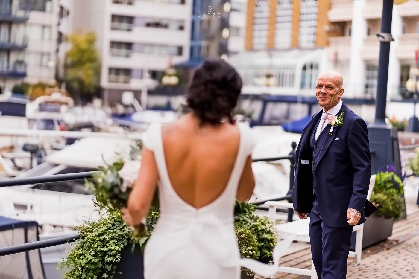 Bride seen from behind in a low-backed dress walks towards the groom, who smiles in a navy three-piece suit beside the moored boats of a marina.