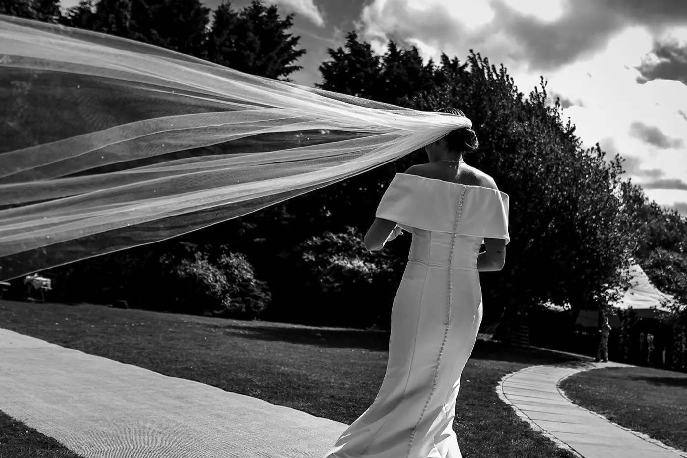 Black and white of a bride from behind walking along a garden path, her long veil blown out sideways by the wind.