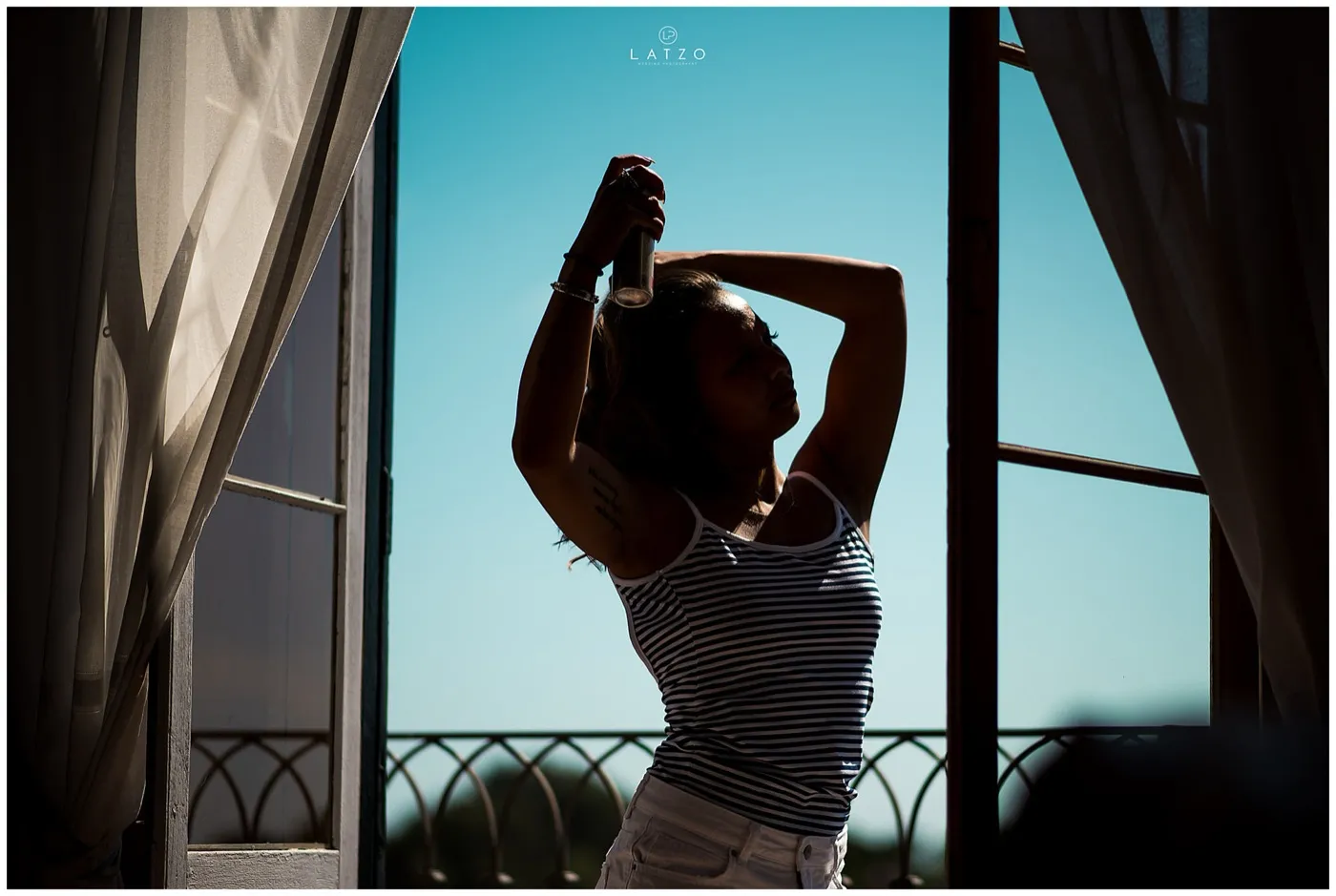 Silhouette of a woman in a striped top spraying her hair in front of an open balcony door, blue sky outside.
