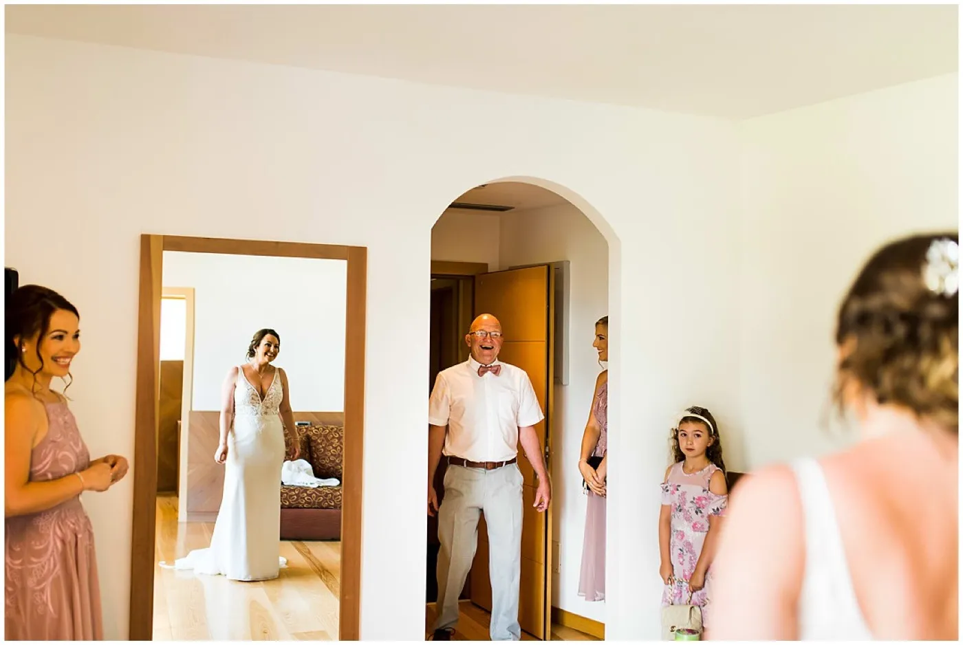 An older man in a pink bow tie laughs in a doorway as the bride, reflected in a mirror, stands in her dress and a flower girl watches.