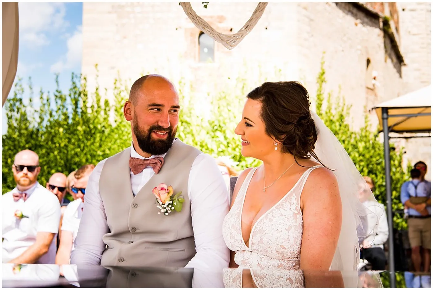 Groom in a grey waistcoat and pink bow tie smiles at the bride during the ceremony at the castle, guests in sunglasses sitting behind them.