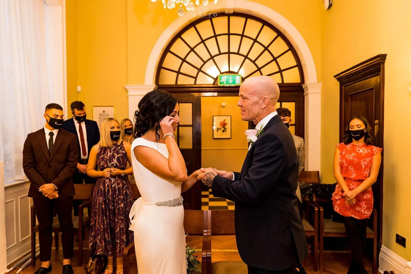 Bride wipes away a tear as she holds the groom’s hands at the ceremony in a yellow room with an arched window, family in black face masks standing behind.