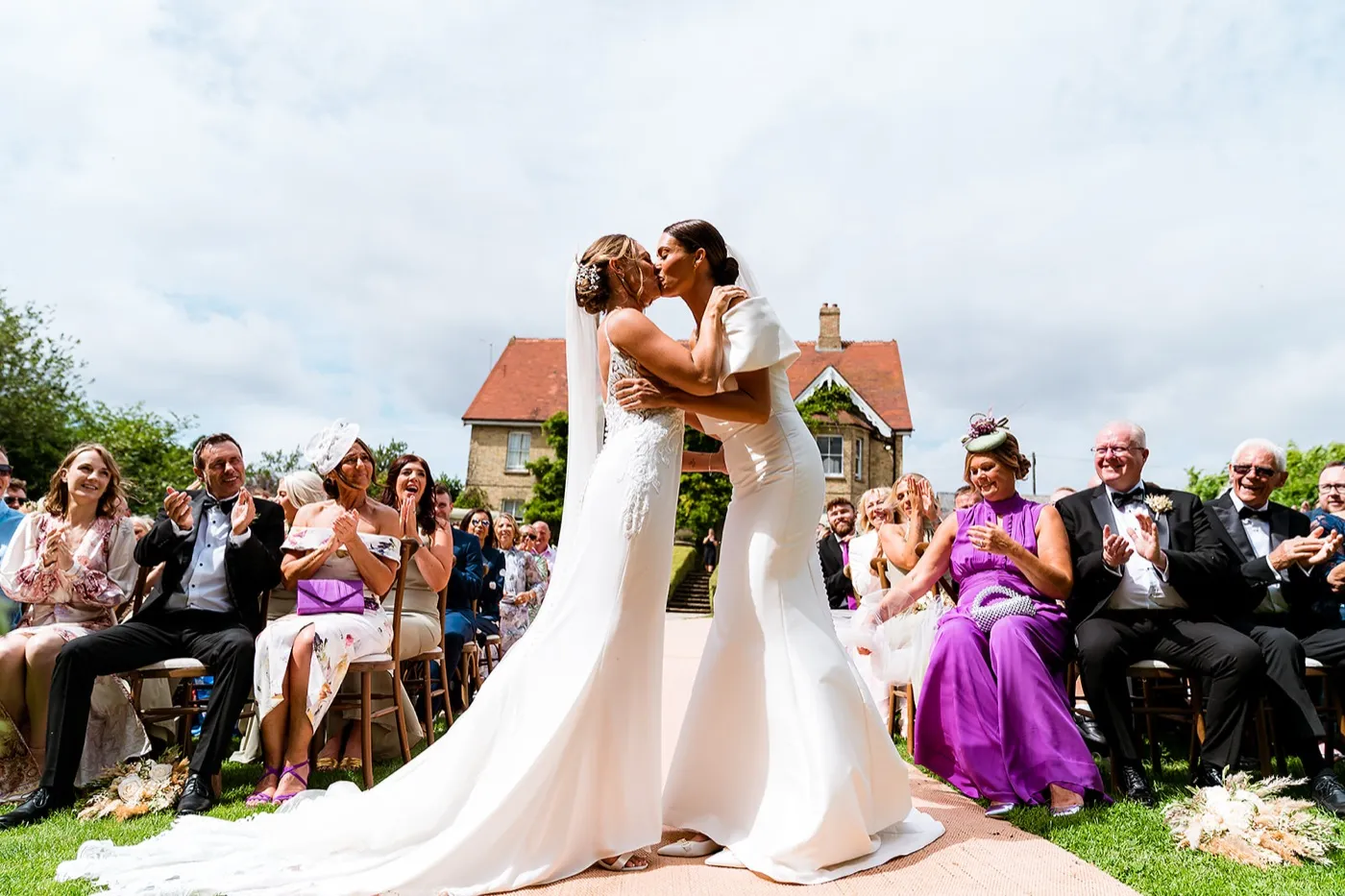 Two brides kiss at the end of the aisle on the lawn as seated guests clap and cheer, the farmhouse behind them.