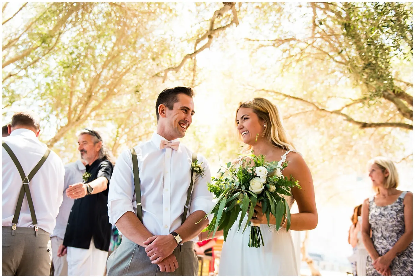 Groom in braces and a pink bow tie laughs with the bride, who holds a bouquet of white roses, under the branches of olive trees.