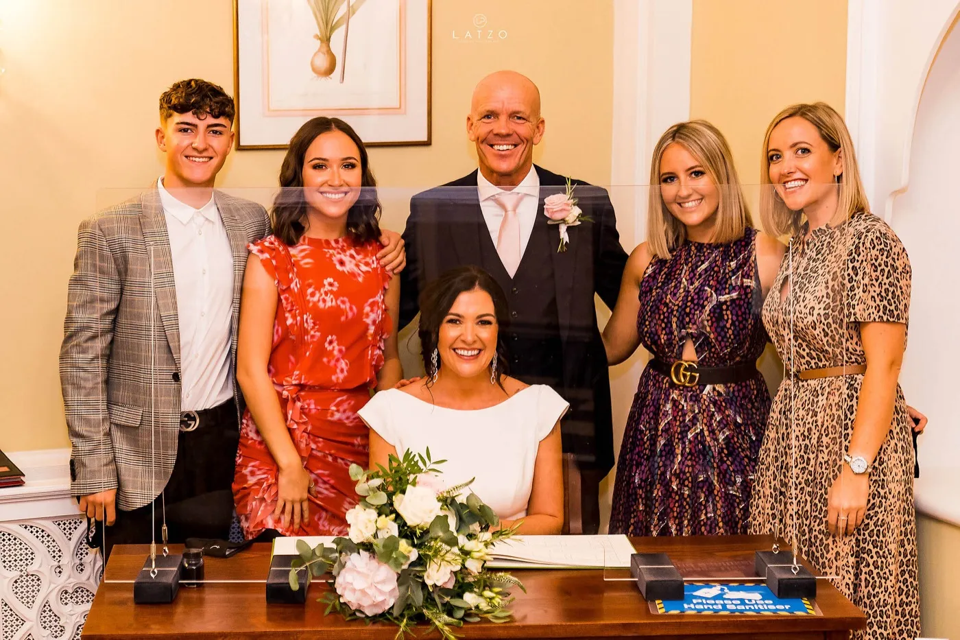 Bride sits smiling at the signing table behind a clear screen, the groom and four young adults standing around her.