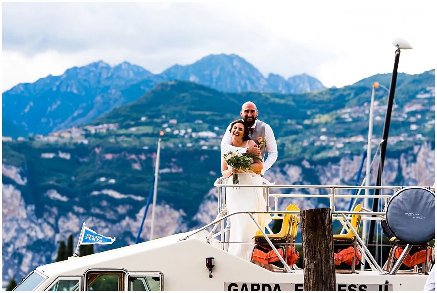 Groom hugs the laughing bride from behind on the front deck of a boat, green mountains and hillside villages rising behind them.