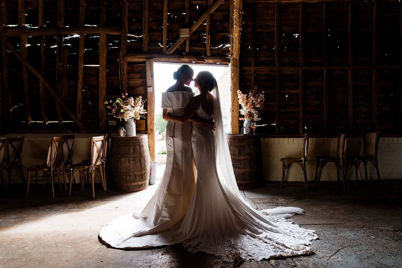 Two brides hold each other in the doorway of a dark timber barn, backlit, their long trains spread across the floor.