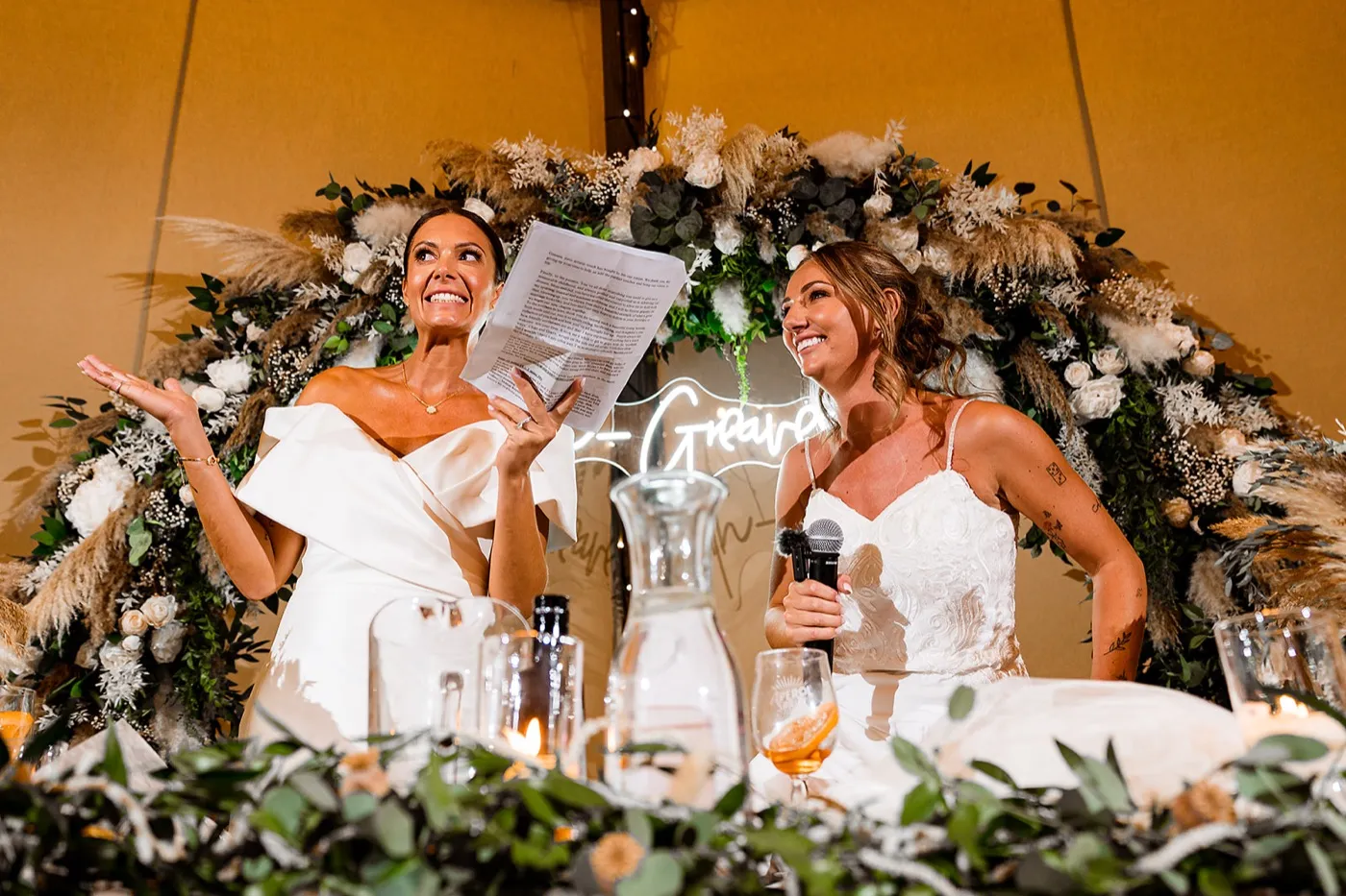 Two brides laugh at the top table in front of a dried flower arch, one holding her speech notes and the other a microphone.