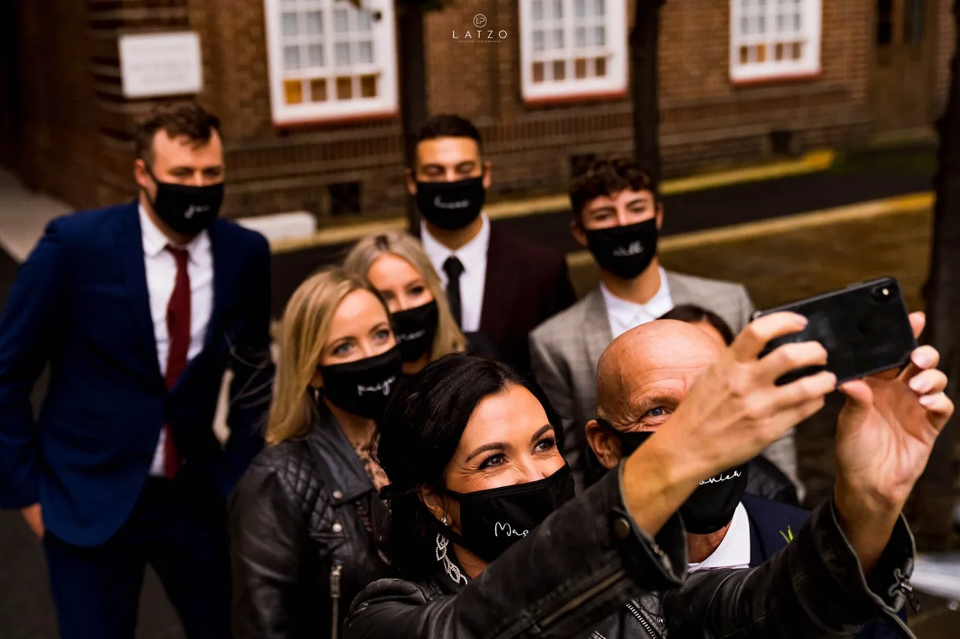 Bride in a leather jacket takes a selfie with the groom and family, everyone in black face masks with names written on them.