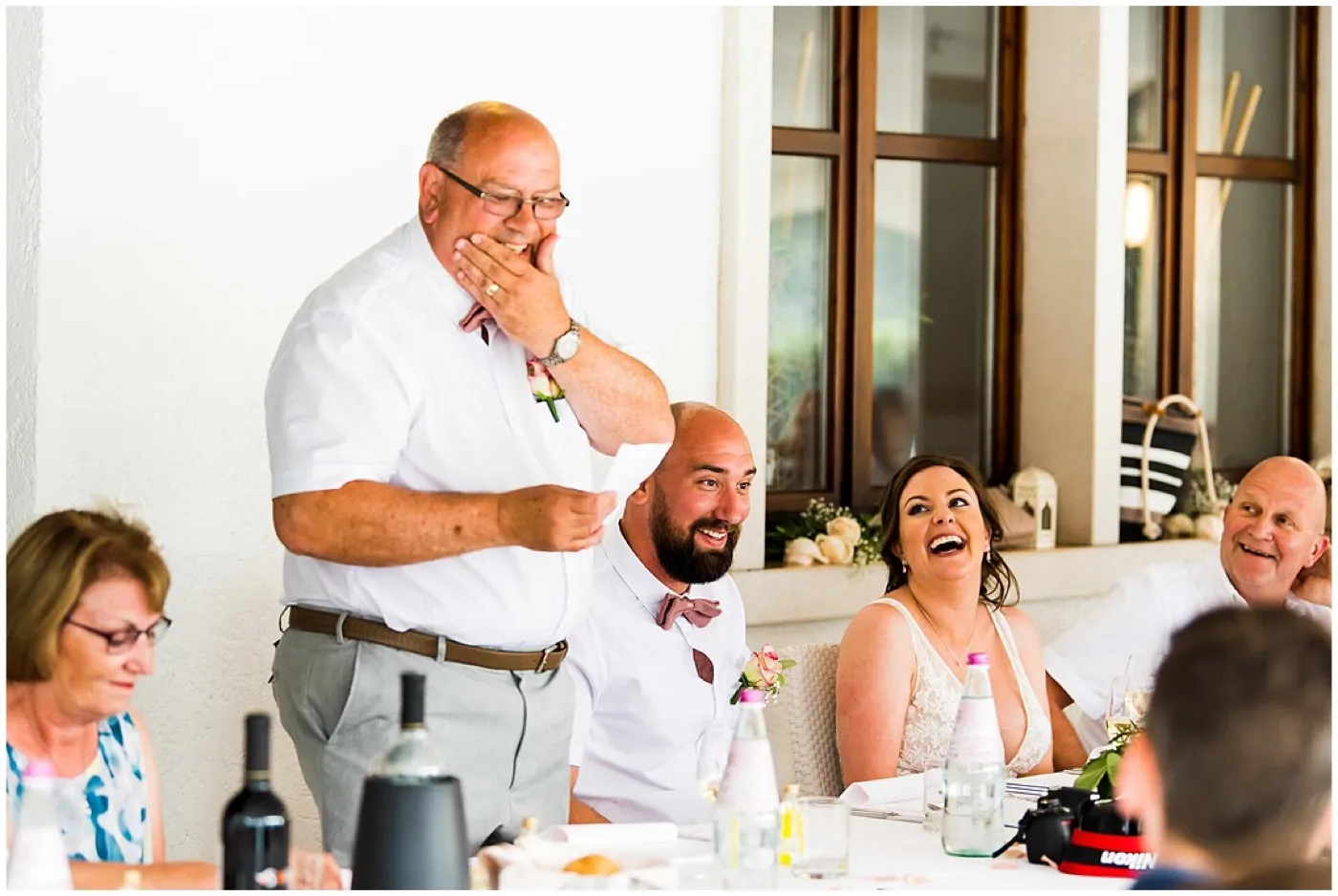 An older man in a white shirt covers his mouth mid-speech as the bride and groom laugh beside him at the reception table.