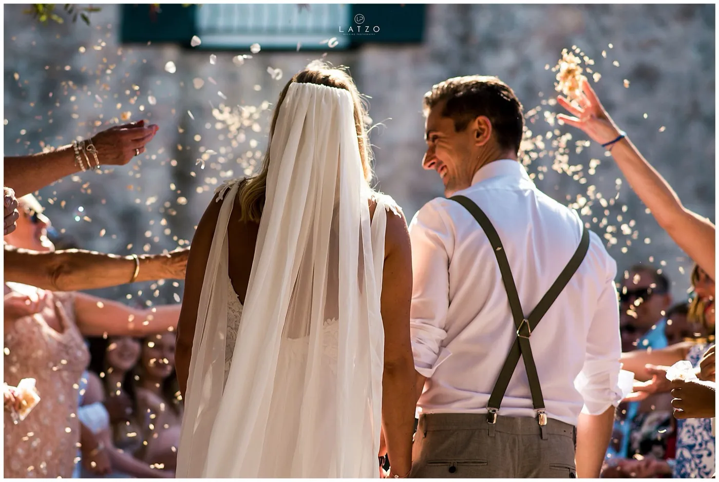 Guests throw confetti over the bride and groom as they walk away hand in hand past an old stone wall, the groom grinning back over his shoulder.