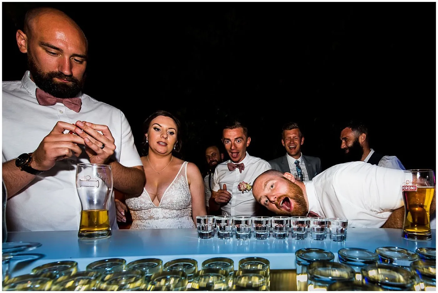 Groom fiddles with his wedding ring at a bar lined with shot glasses as the bride watches and a guest rests his head on the bar, mouth wide open.