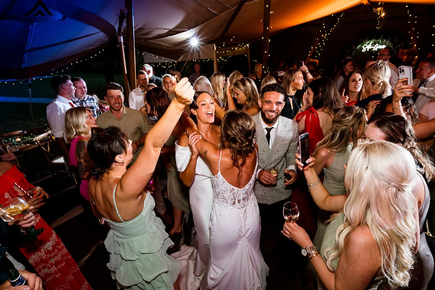 Two brides dance in the middle of a packed tipi as friends cheer, film on their phones and raise their arms around them.