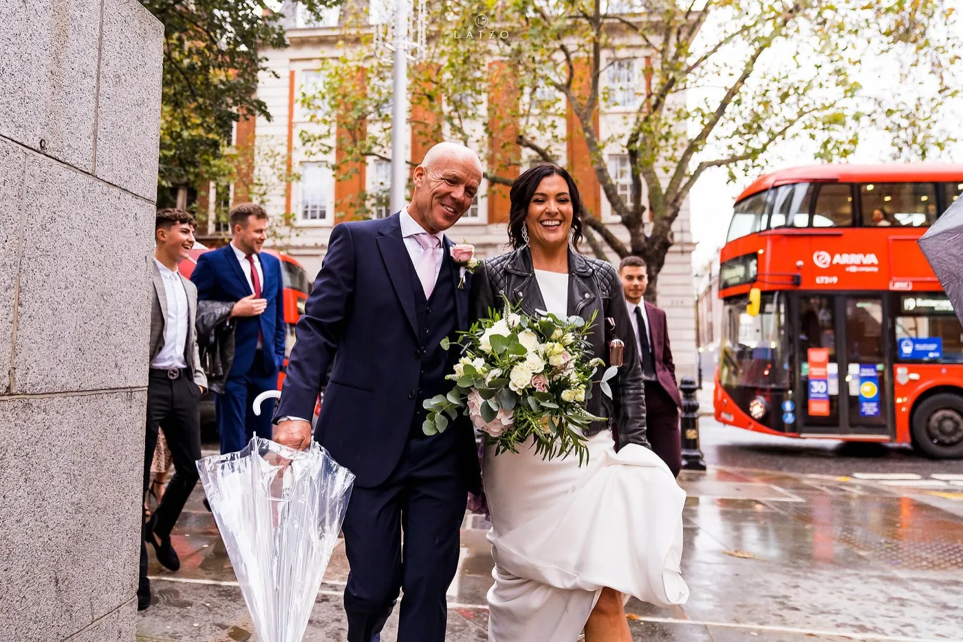 Bride in a leather jacket and groom with a clear umbrella laugh as they walk along a wet London street, a red double-decker bus behind them.