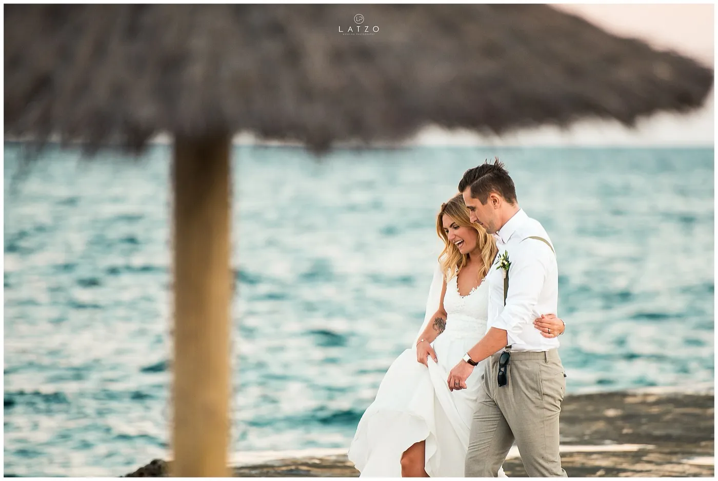 Bride and groom walk arm in arm along the rocky seafront, laughing, a thatched beach umbrella out of focus in front of them.