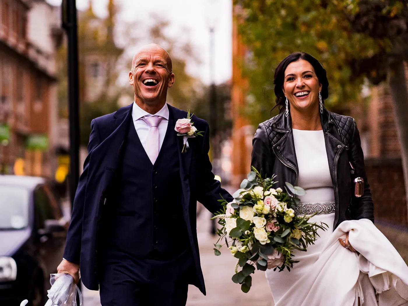 Groom laughs with his mouth wide open as he walks hand in hand with the smiling bride along a tree-lined London pavement.