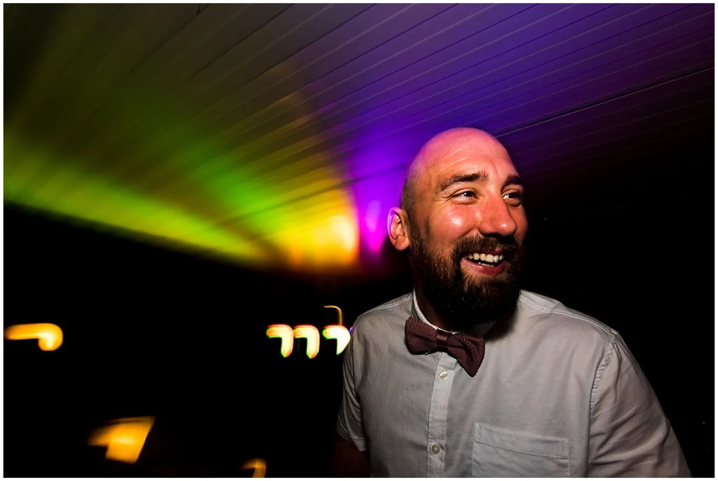 Groom in a bow tie laughs under green, yellow and purple party lights on the dance floor.
