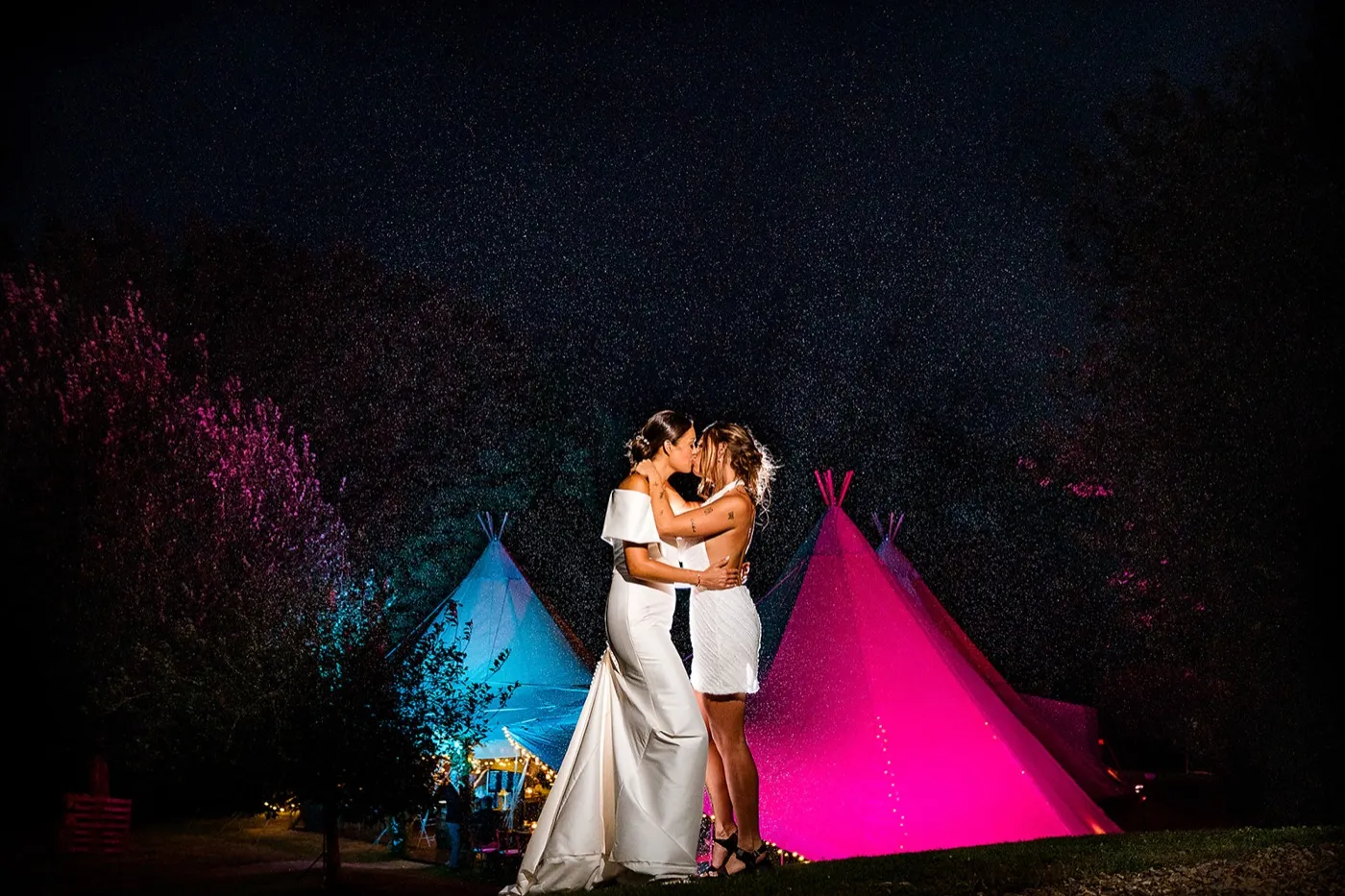 Two brides kiss at night in front of tipis lit pink and blue, the dark sky speckled with light.