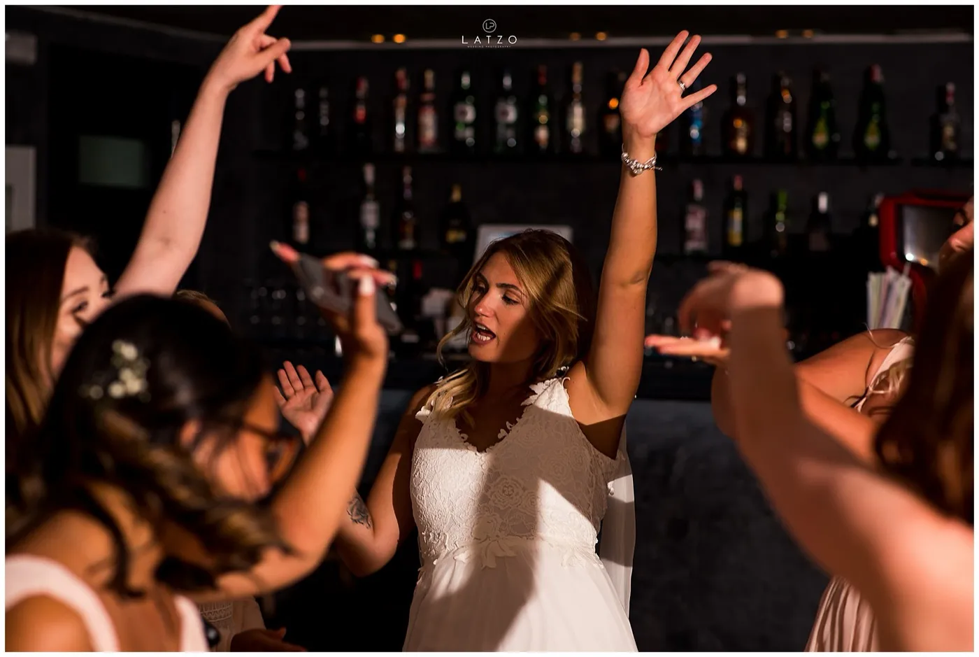 Bride dances with her arms up among her friends in front of a bar lined with bottles.