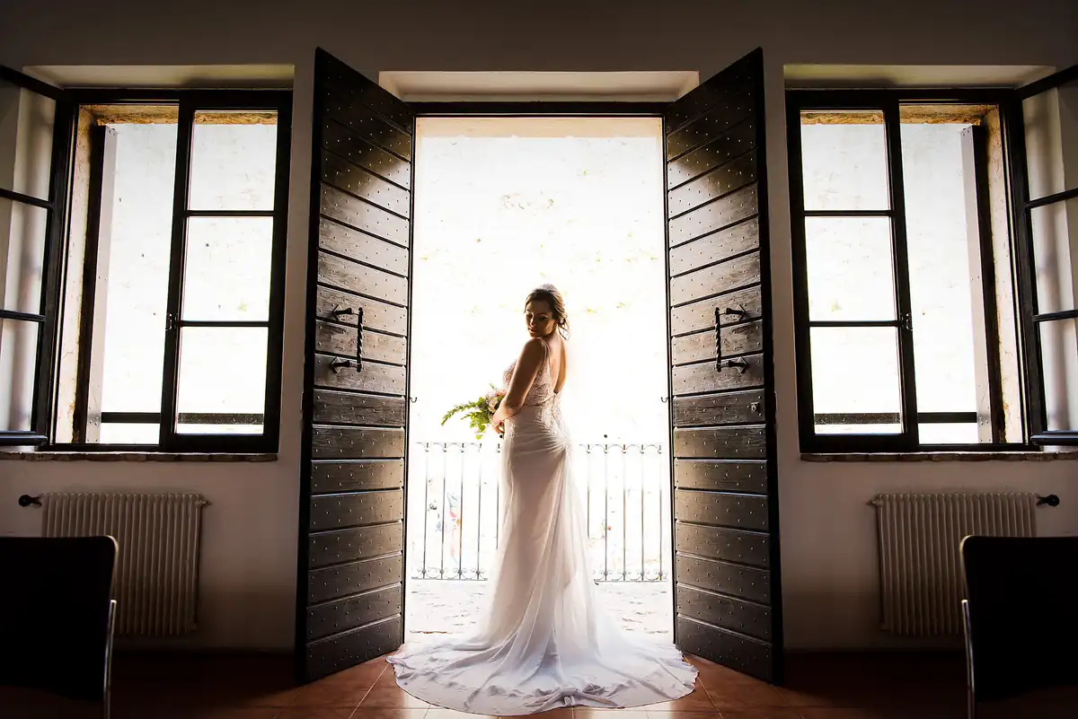 Bride stands in a tall open doorway between dark wooden doors, lit from behind by a bright balcony.