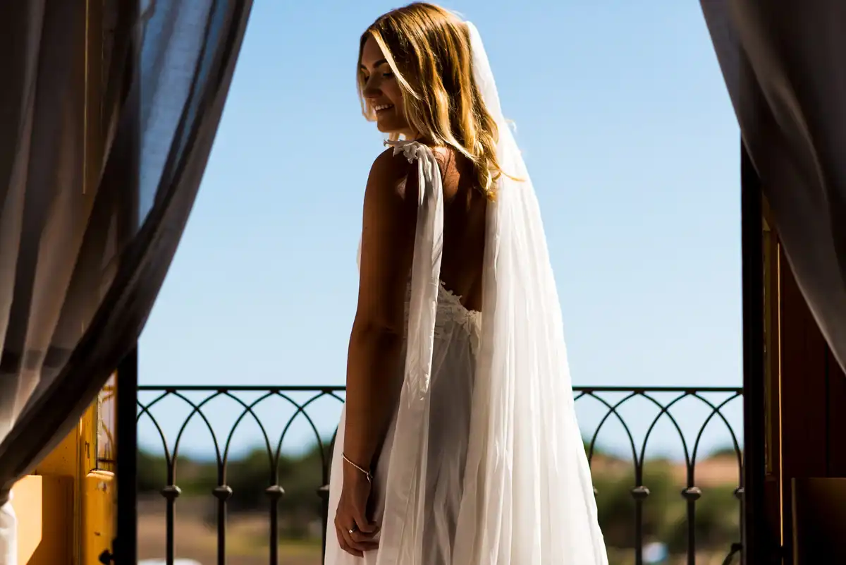 Bride in a long veil looks down and smiles on a sunny balcony, framed by curtains and blue sky.