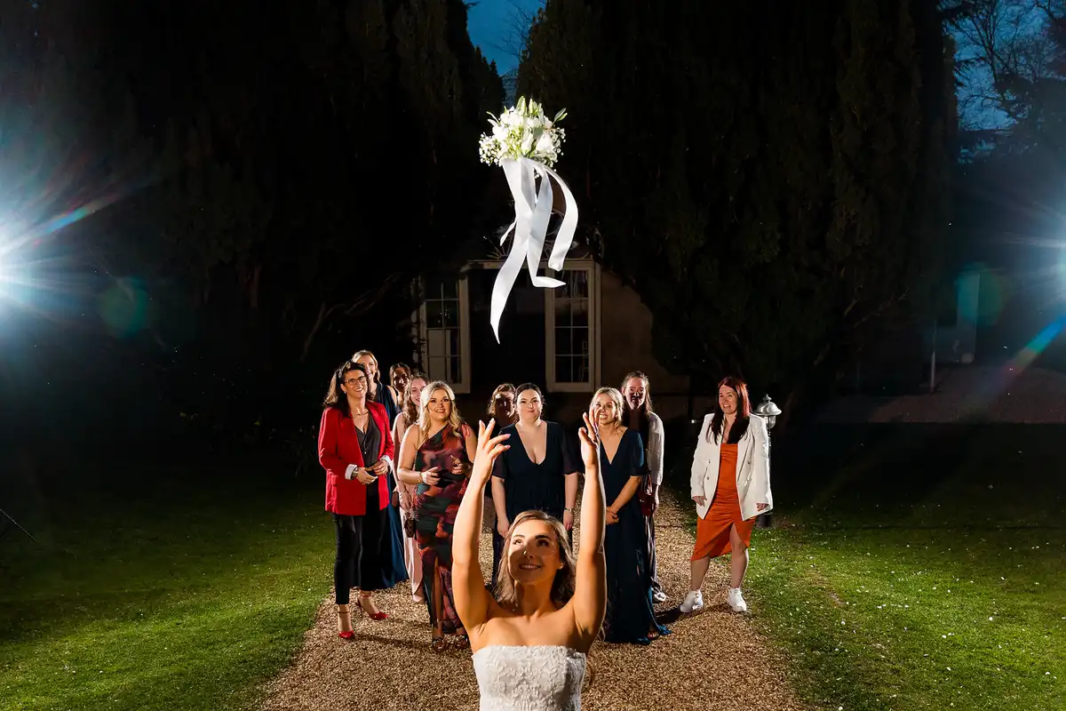 Bride tosses her bouquet back over her head towards a line of guests on a gravel path at night.