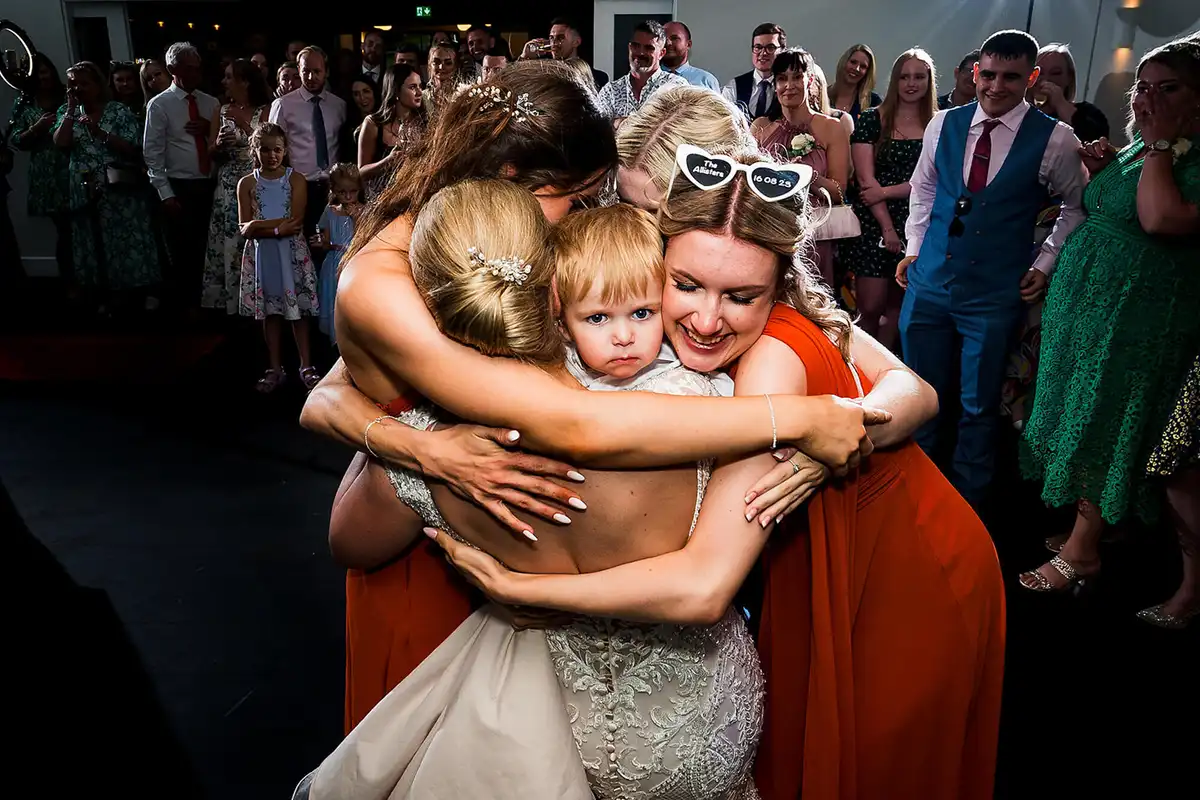 Bride and bridesmaids in orange wrap a toddler in a group hug on the dance floor as guests watch.