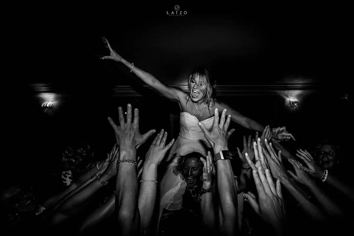 Black and white of the bride crowd surfing on a sea of raised hands, one arm flung out, grinning.
