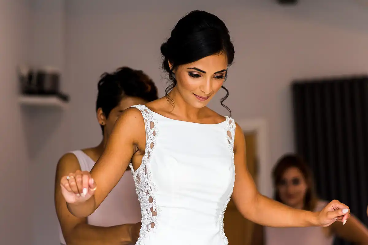 Bride dances with her arms out as a bridesmaid fastens the back of her dress, another smiling behind.