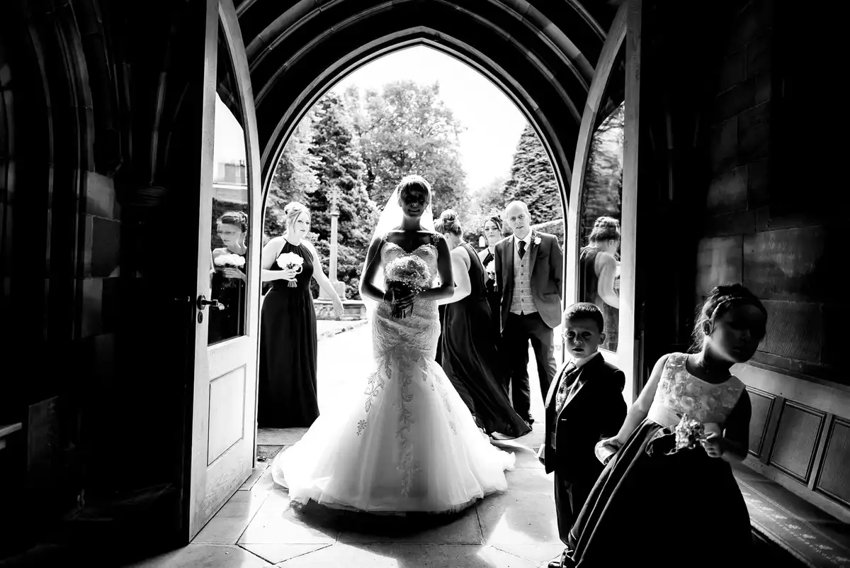 Black and white of the bride walking into a church through a pointed arched doorway, a flower girl and page boy in shadow.