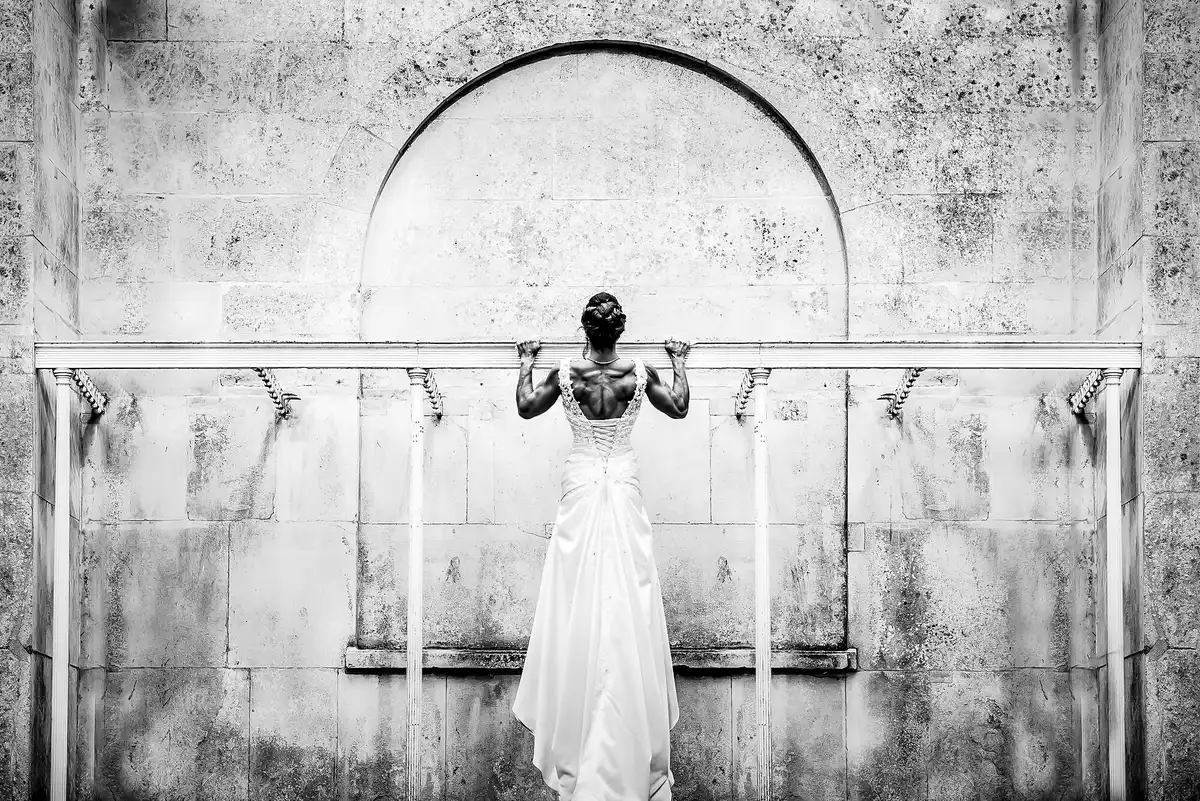 Black and white of the bride from behind, gripping a metal rail on a pale stone wall beneath an arch.