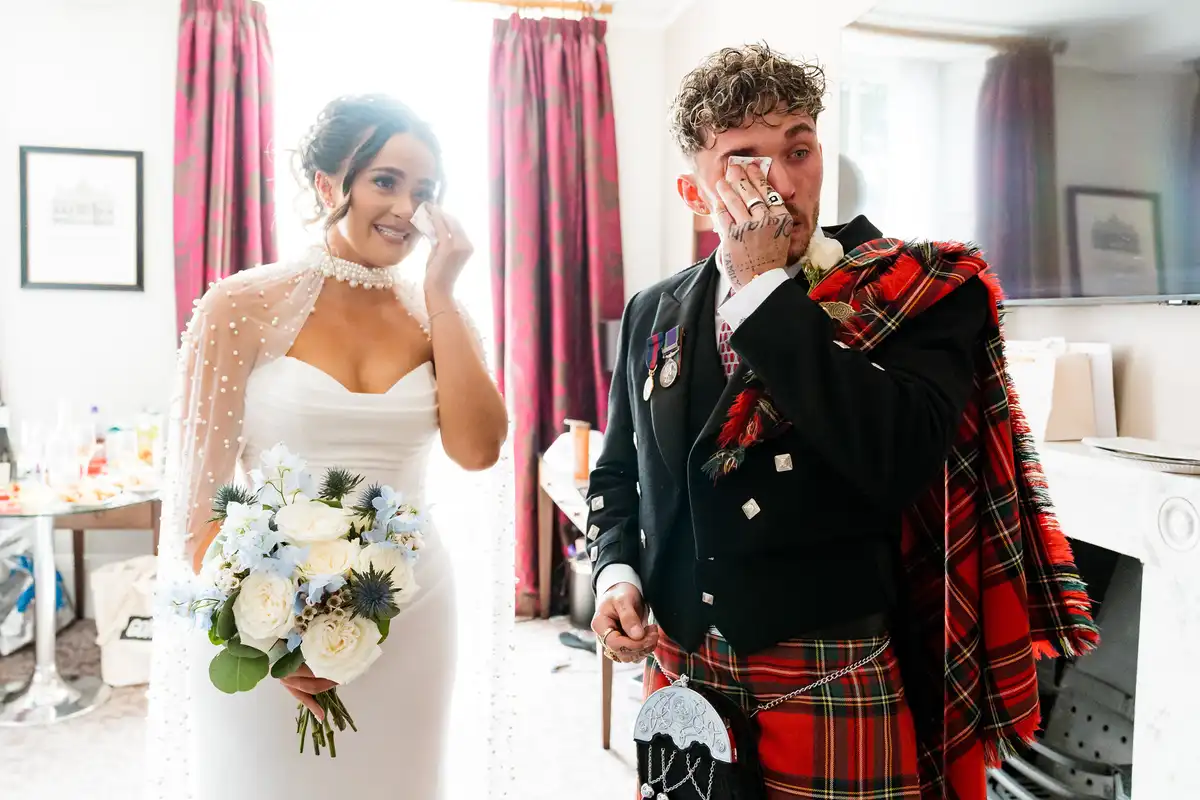 Bride and groom in a tartan kilt both wiping away tears in a hotel room at their first look.