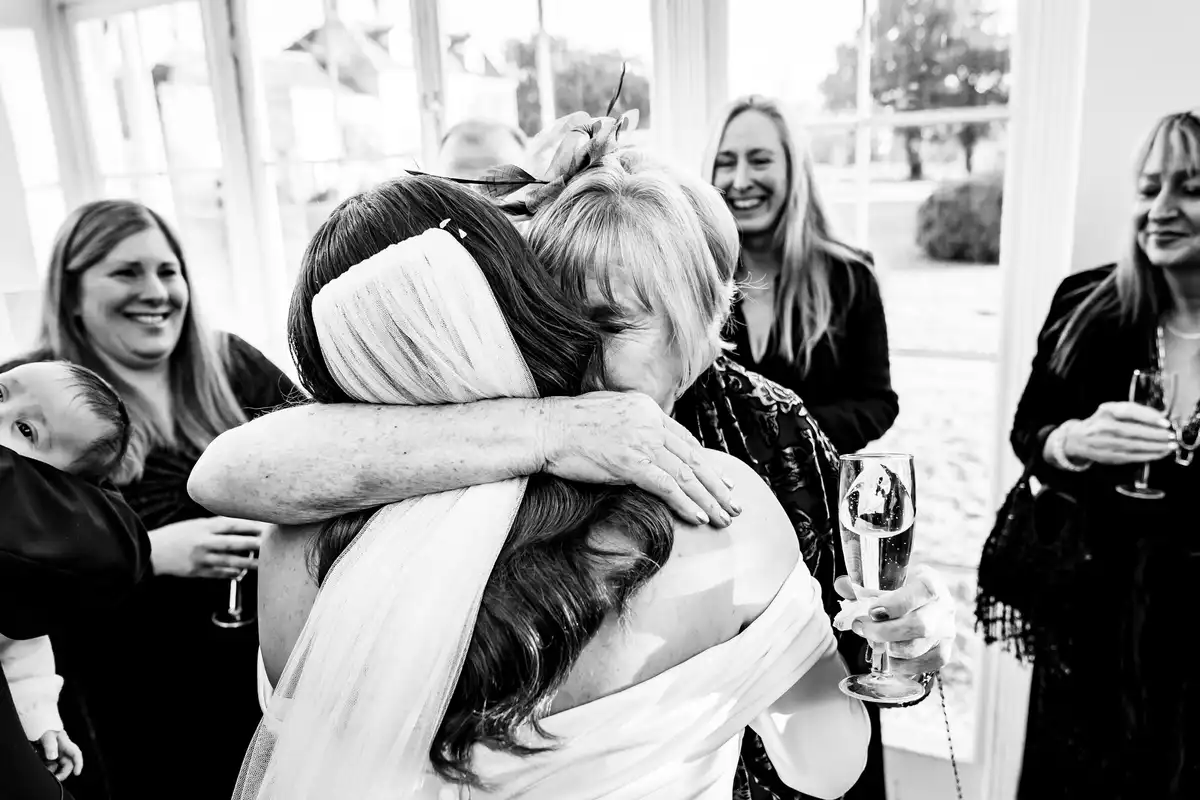 Black and white of the bride in a tight hug with an older woman as smiling guests look on.