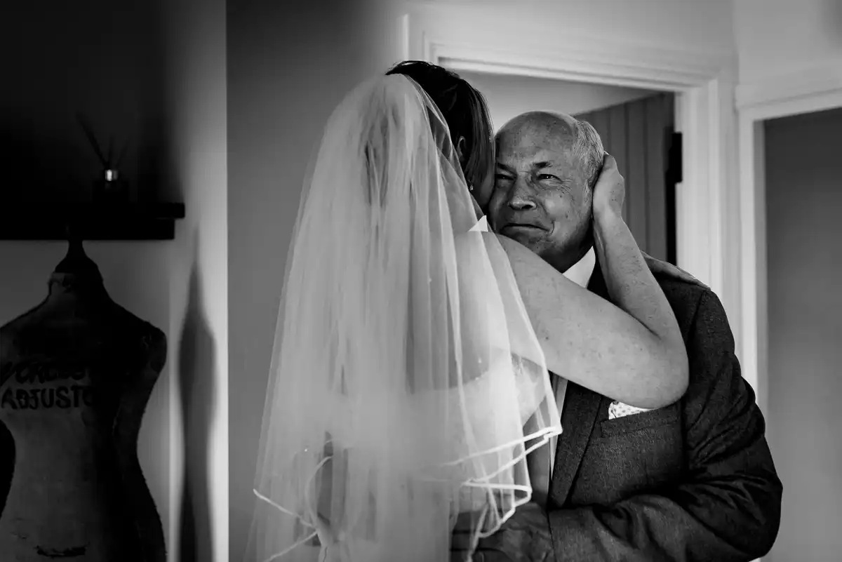 Black and white of the bride in her veil hugging an older man in a doorway, a dressmaker's dummy beside them.