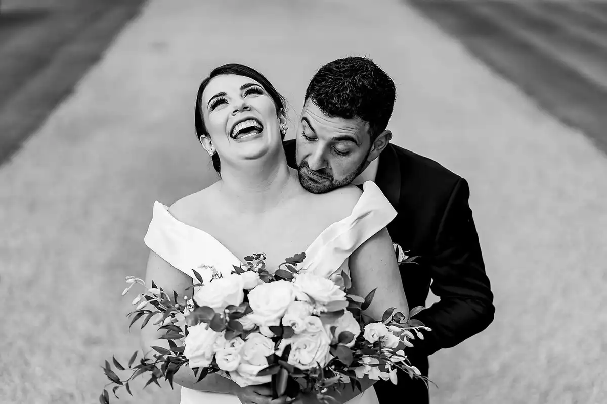 Black and white of the bride laughing, head tipped back, as the groom rests his face on her shoulder.