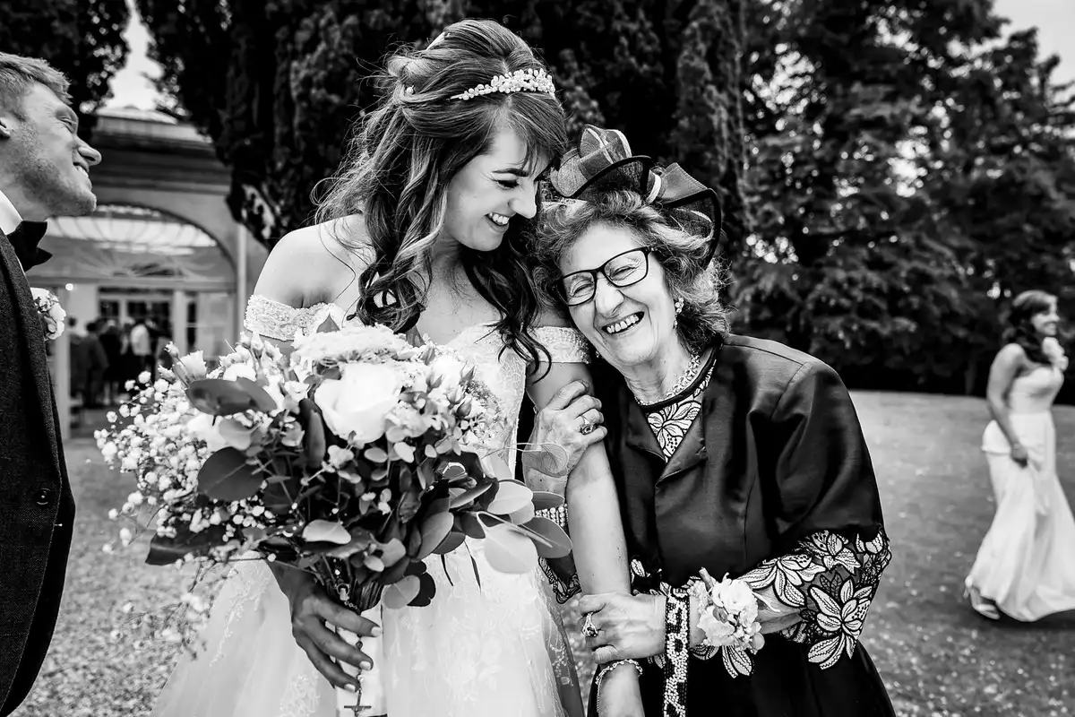 Black and white of the bride laughing, arm in arm with an older woman in glasses and a fascinator.