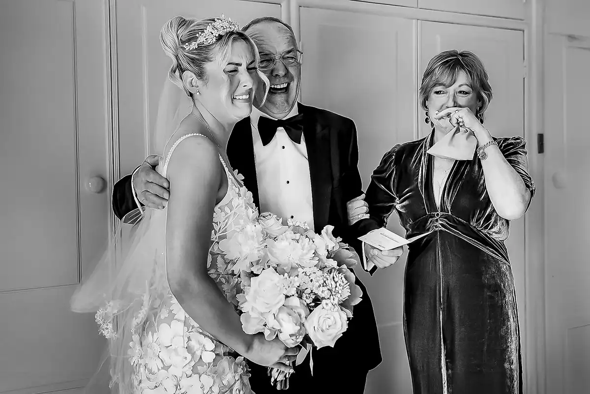 Black and white of a tearful bride hugged by a laughing older man, an older woman crying beside them.
