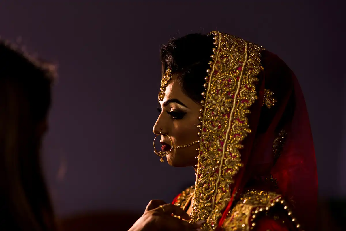 Side view of a bride in a red and gold embroidered dupatta and nose ring against a dark purple background.