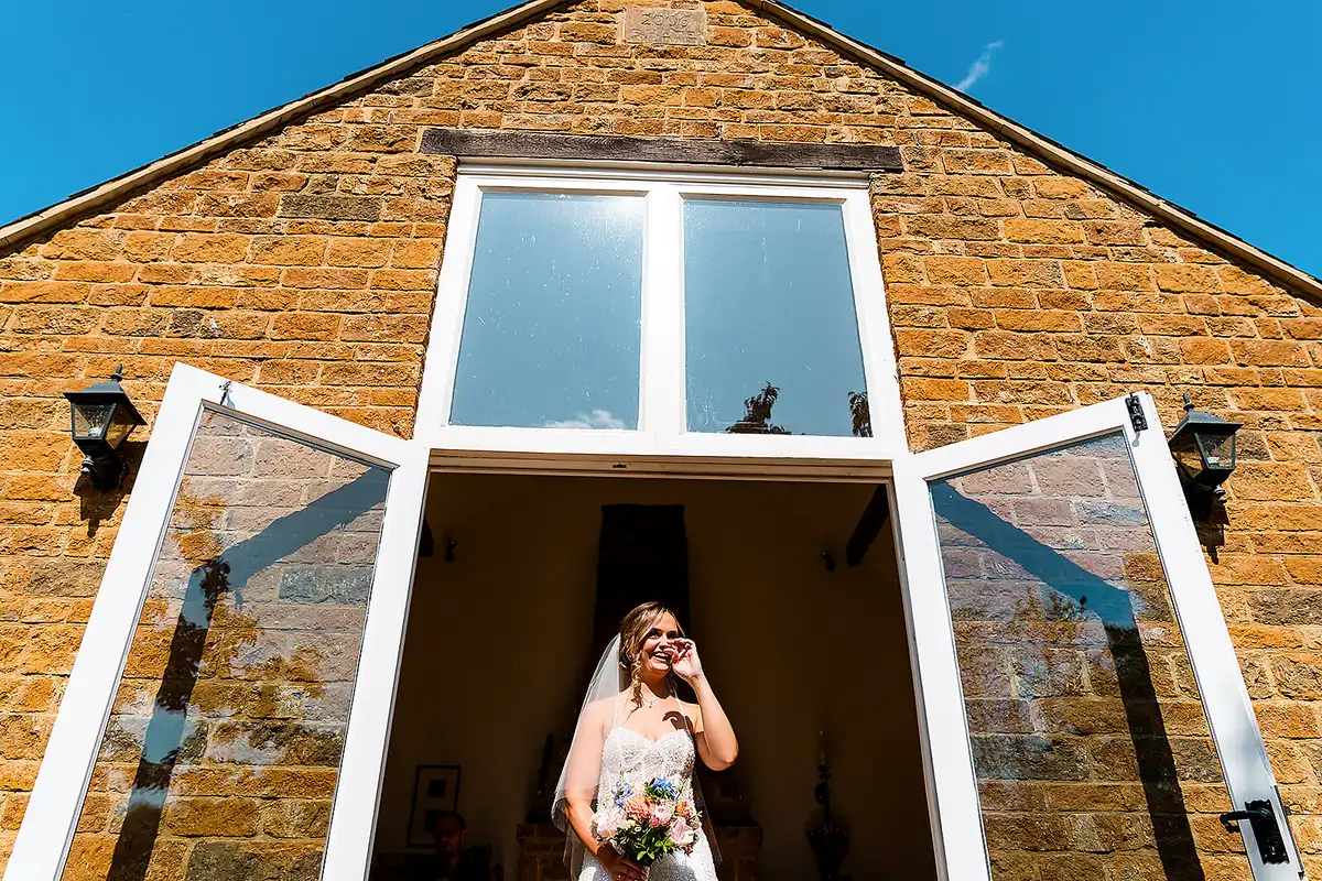 Bride holding her bouquet wipes away a tear in the open glass doors of a stone barn under a blue sky.