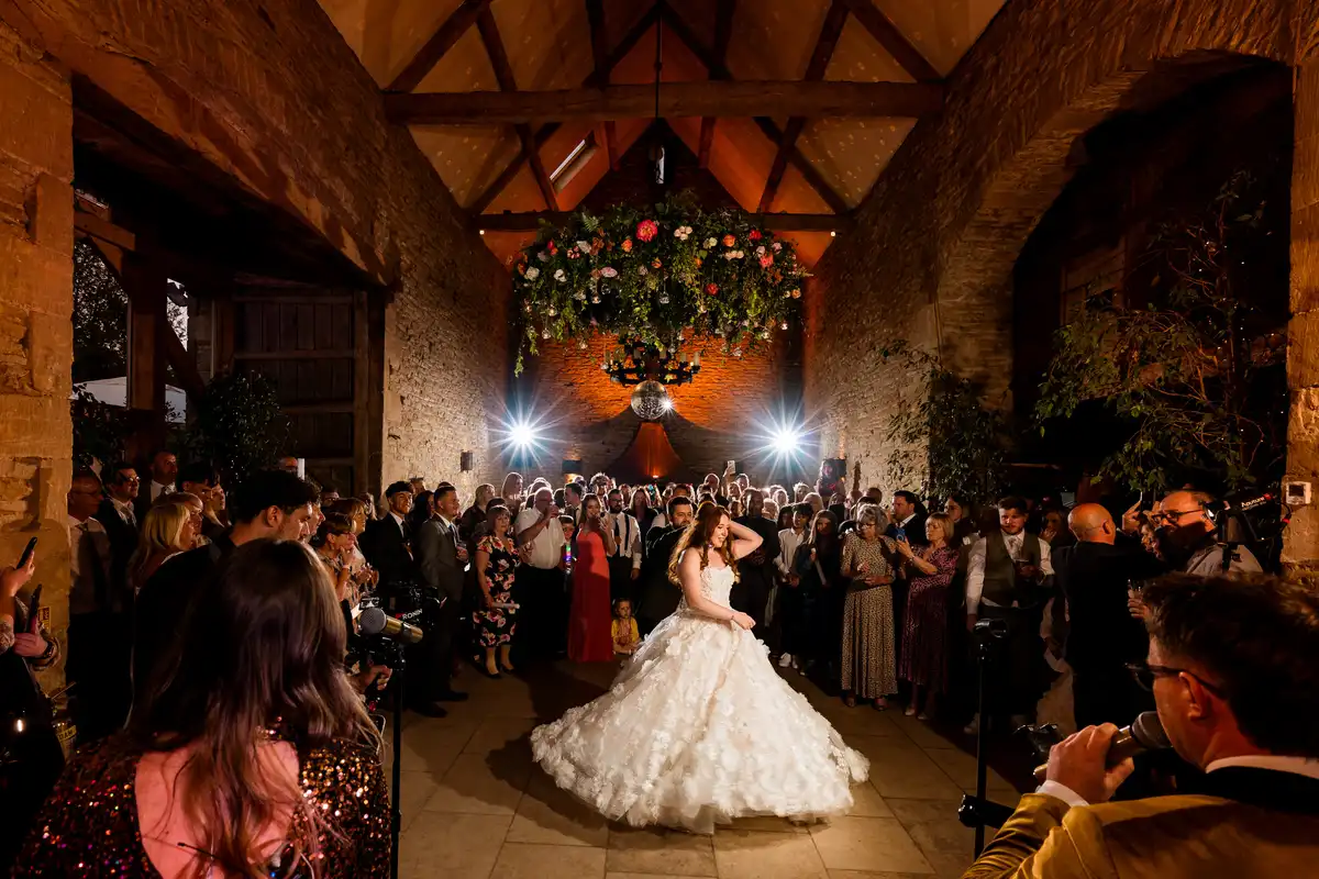 Bride twirls her full skirt in a stone barn as guests circle round and a singer performs.