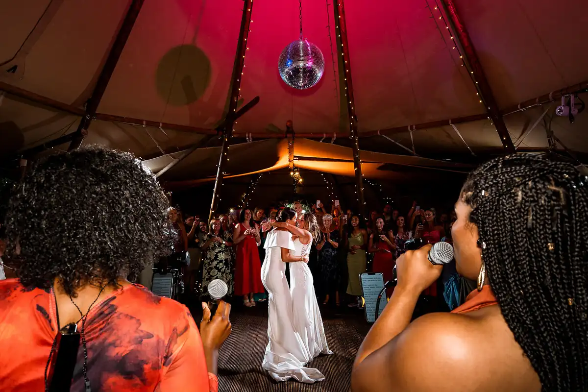 Two brides share their first dance in a tipi under a disco ball as friends sing into microphones.