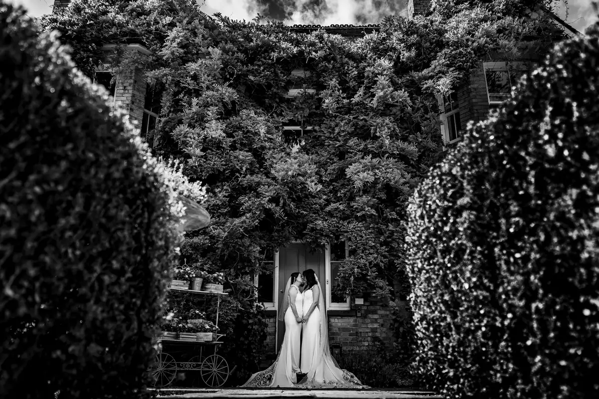 Black and white of two brides kissing at the door of an ivy-covered house, framed by tall hedges.