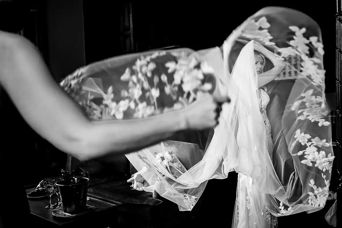 Black and white of a bridesmaid flinging a long lace veil out over the bride as she gets ready.